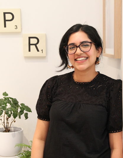 A young woman wearing glasses stands next to a plant. Two letters on the wall int  he background spell out "PR".